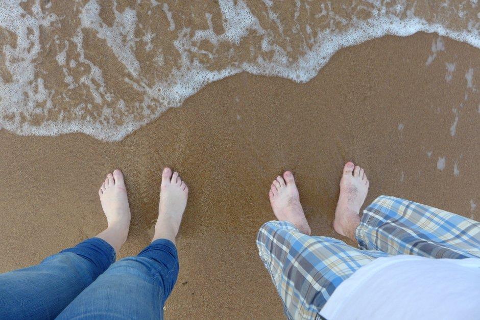 Two pairs of feet standing on sand, small wave coming up to meet them