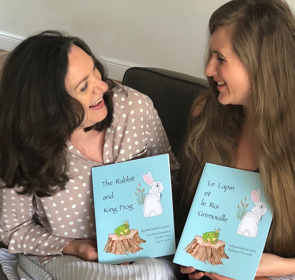 Patti and her daughter Marianne hold French and English copies of a children's book: The Rabbit and King Frog. They are looking at each other and smiling